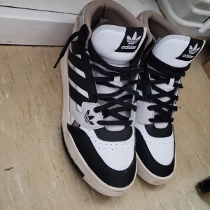 Adidas sneakers med svartvita detaljer - Säljer ett par Adidas sneakers i svart, vitt och beige med klassiska tre ränder på sidorna. Skorna har snörning, hög modell och Adidas logga på plösen. Perfekta för dig som gillar streetwear och sportig stil.