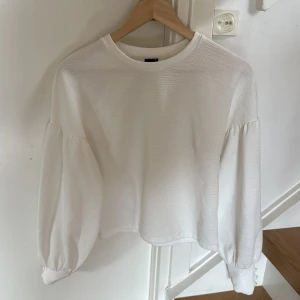 Vit långärmad topp från Gina Tricot - Säljer en vit långärmad topp från Gina Tricot i storlek XS. Tröjan har ballongärmar med knappar vid ärmslut och en lätt croppad passform. Perfekt till lager-på-lager eller som den är.