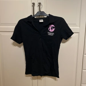 Svart pikétröja från Tee Jays med Travel Shop-tryck - Svart vintage pikétröja från Tee Jays med rosa och vitt Travel Shop-tryck på bröstet. Klassisk krage och knappar framtill. I strl L men sitter bra på S o M 