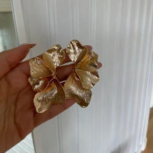 Stora blommiga örhängen i guld oanvända  - Säljer ett par stora, blommiga örhängen i guldmetall. De har en snygg, organisk form som påminner om blomblad och ger ett statement-intryck. Perfekta för dig som vill sticka ut med dina accessoarer.