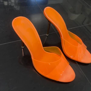 Orange klackskor med öppen tå - Säljer ett par trendiga orange klackskor med öppen tå och transparent detalj över foten. Skorna har hög smal klack och en stilren design som verkligen sticker ut. Perfekta för dig som vill ha något färgstarkt och modernt.