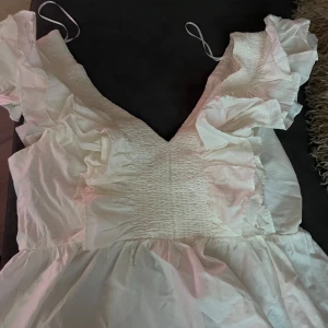 Vit volangtopp från H&M - Den trendiga söta vita toppen från H&M med volangdetaljer och smockad midja. Klänningen har korta ärmar med volanger och en v-ringad hals. Perfekt för sommarens alla tillfällen! Ny med prislapp kvar. 