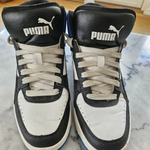 Puma sneakers i svart, vitt och blått - Säljer ett par Puma sneakers i svart, vitt och blått. Skorna har klassisk snörning, rund tå och en platt sula. 