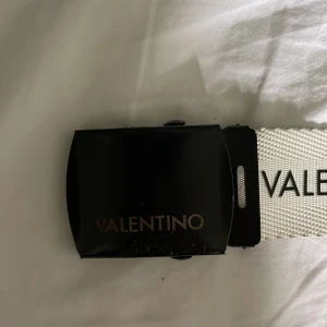 Vitt bälte från Valentino - Säljer ett vitt bälte från Valentino med svart logotyp tryckt längs hela bältet. Bältet har en modern och stilren design med en bred, svart spänne. Perfekt accessoar för att lyfta din outfit. Säljs för 200 kr här av er vid minsta fundering 