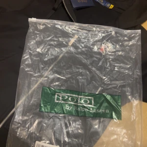 Polo Ralph Lauren   - Svart hoodie med dragkedja från Polo Ralph Lauren. Klassisk design med röd broderad logga på bröstet, huva med dragsko och mjukt innerfoder. Perfekt för en avslappnad och stilren look.