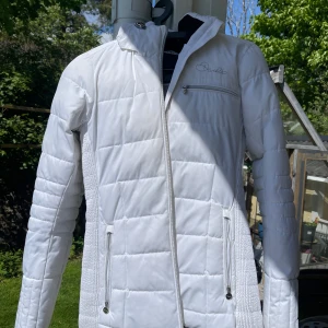 Vit quiltad jacka  - Säljer en snygg vit quiltad jacka med dragkedja framtill och på fickorna. Jackan har lång ärm, hög krage och diskret logga på bröstet. Perfekt för kyligare dagar och ger en sportig look. Har använts som skidjacka