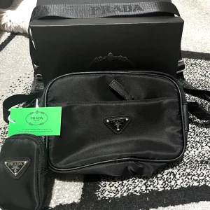 Svart axelväska  med triangel logo - Snygg svart axelväska i syntetmaterial med justerbar axelrem och tillhörande liten väska. Klassisk trekantig logga i metall framtill. Perfekt för att bära dina viktigaste saker med stil.