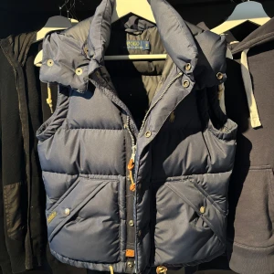 Mörkblå dunväst från Polo Ralph Lauren - Snygg mörkblå dunväst från Polo Ralph Lauren med huva och dragkedja. Västen har tryckknappar, snedställda fickor och klassisk logga på bröstet. Perfekt för lager-på-lager och stilren look. Storlek S men sitter som XS