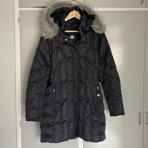 Svart quiltad dunjacka Luhta 40 - Luhta storlek 40. Svart quiltad dunjacka med dragkedja och pälsdetaljer på huvan. Markerad midja, justeras med knapp. Saknas en flärp på en av dragkedjorna på fickorna, syns på bild. 
