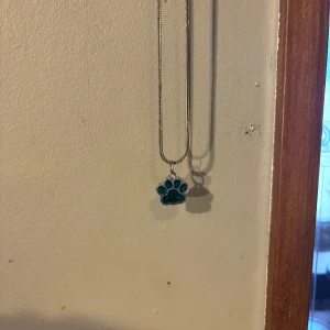 Halsband med tassberlock  - Säljer ett halsband med en silverfärgad kedja och en berlock formad som en tass. Perfekt för dig som älskar djur eller vill ha något unikt.