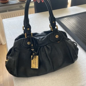 Svart handväska från Marc by Marc Jacobs - Säljer en svart handväska från Marc by Marc Jacobs med guldiga metalldetaljer och dubbla handtag. Väskan har ett innerfoder med bokstavsmönster och dragkedja upptill. Perfekt storlek för vardagens alla prylar.