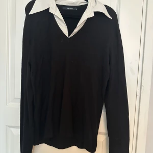 Svart tröja med vit krage från Vero Moda - Snygg svart tröja från Vero Moda med vit skjortkrage och v-ringning. Perfekt för en preppy look. Tröjan har långa ärmar och är tillverkad i ett mjukt syntetmaterial. Klassisk och stilren design.