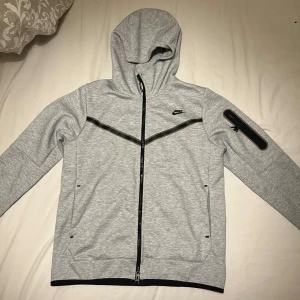 Nike tech hoodie grå - Snygg ljusgrå hoodiejacka från Nike med svarta detaljer och dragkedjor. Jackan har huva, två fickor framtill och en extra ficka med dragkedja på ärmen. Perfekt för en sportig och avslappnad stil.