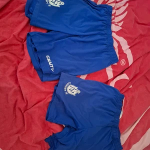 2st Blå träningsshorts från Craft - Säljer flera par blå träningsshorts från Craft med klubbtryck på ena benet. Shortsen har resår i midjan och är tillverkade i ett lätt och luftigt material som passar perfekt för sport och aktivitet.