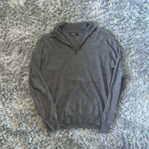 Grå halvzip-tröja från J.Lindeberg - Säljer en stilren grå tröja från J.Lindeberg med halv dragkedja och diskret logga på bröstet. Tröjan är tillverkad i mjuk merinoull och har långa ärmar samt ribbade muddar. Perfekt för lager-på-lager och snygg till både jeans och chinos.