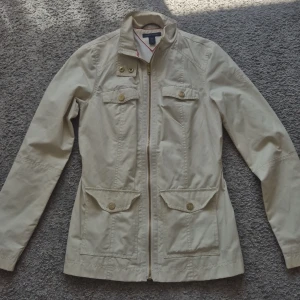 Beige jacka från Tommy Hilfiger - Snygg beige jacka från Tommy Hilfiger med dragkedja och fyra fickor framtill. Jackan har lång ärm, markerad midja och klassisk krage. Perfekt för vår och höst, tillverkad i bomull med detaljer i guldton.