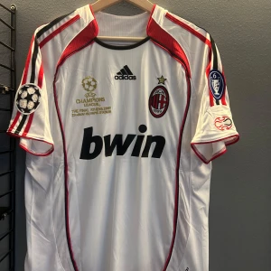 AC Milan fotbollströja Kaka 22 Champions League - Säljer en vit AC Milan fotbollströja med röda och svarta detaljer, Champions League-märken på ärmarna och Kaka 22 på ryggen. Adidas-logga och Bwin sponsortryck på bröstet. Perfekt för fans av italiensk fotboll. En replika helt ny med tagg