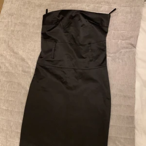 Svart axelbandslös klänning från LiluMary - Säljer en svart axelbandslös klänning från LiluMary med figurnära passform och slits baktill. Klänningen har en elegant och stilren design, perfekt för speciella tillfällen. Materialet känns mjukt och har en lätt glans.