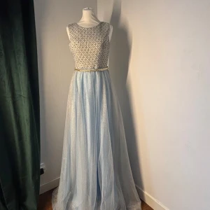 Ljusblå långklänning med glittrig topp och guld-detaljer - Säljer en elegant ljusblå långklänning med glittrig, mönstrad topp och guldigt bälte i midjan. Klänningen är ärmlös och har en öppen rygg med snygg detalj. Orginalpris: 2500kr