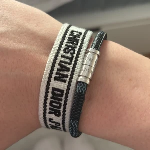 Armband - Två armband: Ett broderat band i vitt och svart med texten 'CHRISTIAN DIOR J'ADIOR' och tofsar, samt ett svart LV armband i flätad syntet med silverfärgat magnetlås. Snygga accessoarer som passar till många stilar. LV armbandet = 19cm, Dior = justerbar passform (Båda för 599jr) -Box ingåt (styckpris = 350kr)