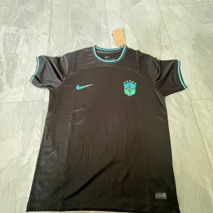 Svart Brasilien fotbollströja från Nike - Säljer en svart Brasilien fotbollströja från Nike med turkosa detaljer och emblem på bröstet. Tröjan har korta ärmar och är tillverkad i Dri-FIT-material som andas. Perfekt för fotbollsträning eller att bära som supporter.