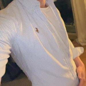 Blårandig skjorta från Ralph Lauren - Säljer en klassisk blårandig skjorta från Ralph Lauren i slim fit-modell. Skjortan har långa ärmar, knappar framtill och den ikoniska broderade loggan på bröstet. Tillverkad i 100% bomull och passar perfekt till många olika tillfällen.lite liten i storleken 