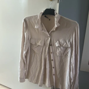 Beige långärmad skjorta med fickor - Säljer en stilren beige skjorta med långa ärmar och två bröstfickor. Skjortan har klassisk krage och knäppning framtill med små knappar. Perfekt att styla till olika outfits för en clean look. Tyvärr lite skrynklig men det är för att den är nytvättad! 