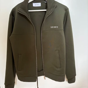 Olivgrön track jacket från Les Deux - Säljer en olivgrön track jacket från Les Deux med dragkedja framtill och ståkrage. Jackan har två fickor på sidorna och diskret logga på bröstet. Perfekt för en sportig och stilren look. Har tyvärr en liten slitning på ryggen men märks knappt, annars i väldigt bra skick
