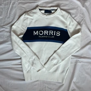 Vit stickad tröja från Morris Rowing Club - Vit stickad tröja från Morris Rowing Club med blått och mörkblått band över bröstet och broderad logga. Rund halsringning och ribbade muddar vid ärmslut och nederkant. Perfekt för en stilren och sportig look.