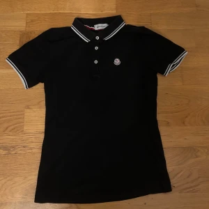 Svart pikétröja från Moncler - Säljer en svart pikétröja från Moncler med vitrandiga detaljer på krage och ärmslut. Tröjan har klassisk pikékrage, knappar framtill och Moncler-logga på bröstet. Perfekt för en stilren och sportig look.