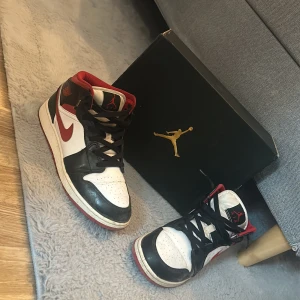 Nike Air Jordan 1 Retro mid OG svart/röd/vit - Säljer ett par klassiska Nike Air Jordan 1 Retro mid OG i svart, rött och vitt. Skorna har högt skaft, röd yttersula, svart snörning och ikoniska Jordan-loggan på plösen. Perfekta för dig som gillar streetwear och sneakerskultur. TVÄTTAS INANN