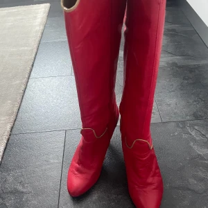 Röda höga stövlar med klack - Säljer ett par riktigt snygga röda stövlar med hög klack och spetsig tå. Stövlarna har dekorativa sömmar i kontrastfärg och går upp till knäna. Perfekta för dig som vill sticka ut med din stil.
