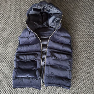 Mörkblå dunväst med huva från Vouter - Säljer en mörkblå dunväst från Vendiera med  huva och dragkedja framtill. Västen har flera stora fickor med lock och en quiltad design som ger en sportig look. Perfekt för lager-på-lager under kyliga och varma dagar