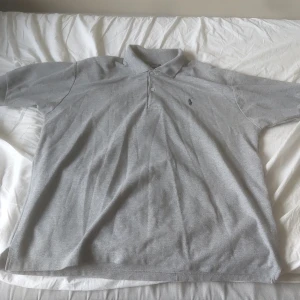Grå pikétröja från Polo - Säljer en klassisk grå pikétröja från Polo i storlek 3XL. Tröjan har korta ärmar, krage med knappar och den ikoniska broderade loggan på bröstet. Tillverkad i 100% bomull för en skön och luftig känsla.