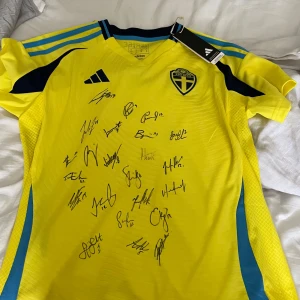 Svensk landslagströja från Adidas - Gul och blå kortärmad fotbollströja från Adidas med Sveriges landslagsemblem på bröstet och flera autografer på framsidan. Tröjan har svarta och blå detaljer på axlar och sidor samt officiell tag kvar. Signerad av Sveriges damlandslag.