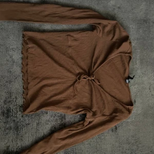 Brun ribbad långärmad topp med knyt - Säljer en brun långärmad topp med ribbad struktur och knyt framtill. Toppen har vågiga kanter nedtill och en smickrande v-ringning. Perfekt för dig som gillar trendiga detaljer och mjuka material.