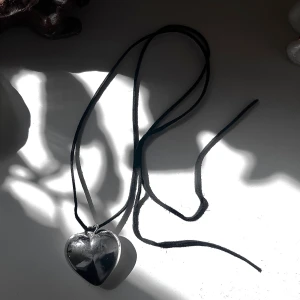 Halsband med silverfärgat hjärta - Poppis och ombloggat i 90-tals-vibe-halsband med ett stort, silverfärgat hjärta i metall och svart mockaband. Hjärtats strl är ca 5x4cm och bandet som kan knytas på olika sätt runt halsen är 107cm. Hjärtat har lite repor. 