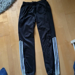 Svarta träningsbyxor med vita ränder - Svarta träningsbyxor med elastisk midja och tre vita ränder längs sidorna på båda benen. Klassisk sportig look, perfekta för gymmet eller fritiden. Byxorna har en smal passform och är tillverkade i syntetmaterial.