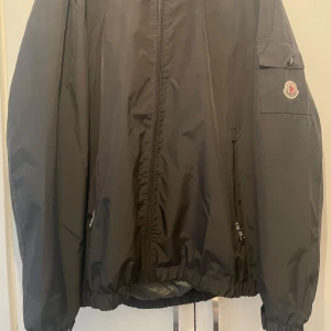 Svart vindjacka från Moncler - Säljer en svart vindjacka från Moncler med luva och dragkedja framtill. Jackan har Moncler-logga på ärmen, fickor med dragkedja och elastiska muddar vid ärmslut och nederkant. Perfekt för blåsiga dagar. Har prislapp men den sitter inte på jackan.