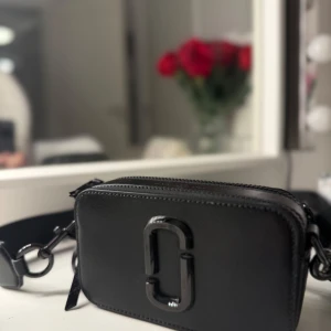 Svart axelväska från Marc Jacobs - Säljer en svart axelväska från Marc Jacobs med bred justerbar axelrem och dekorativ logga framtill. Väskan har dragkedja upptill och är tillverkad i ett tåligt material med stilren design. Perfekt för att bära det viktigaste med stil.