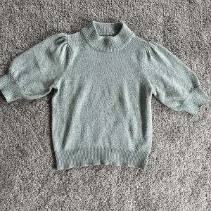 Gröngrå stickad tröja med puffärm - Superfin grön stickad tröja med korta puffärmar och hög ribbad krage. Tröjan har ribbade muddar vid ärmslut och nederkant, vilket ger en snygg passform. Perfekt att styla till jeans eller kjol.