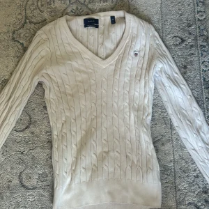 Vit kabelstickad tröja från Gant - Säljer en klassisk vit kabelstickad tröja från Gant med v-ringning och broderad logga på bröstet. Tröjan är i stretchig bomull och har långa ärmar. Perfekt för en stilren och tidlös look.