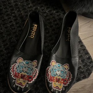 Svarta Kenzo loafers med broderad tiger - Säljer ett par svarta loafers från Kenzo med broderad tiger i färgglada nyanser framtill. Skorna är i skinn och har rund tå samt platt sula. Perfekta för dig som vill sticka ut med en ikonisk detalj från Kenzo.