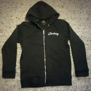 Svart hoodie med dragkedja från Burberry - Säljer en svart hoodie från Burberry med vit broderad logga både fram och bak. Tröjan har dragkedja, huva och långa ärmar. Perfekt för dig som gillar klassisk streetstyle och vill ha något bekvämt och snyggt.