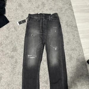 Grå slitna jeans med tapered fit - Säljer ett par grå jeans med slitningar och tvättade detaljer. Byxorna har fem fickor, dragkedja och knapp i midjan. Modellen är tapered och har en modern look med lite smalare ben nertill. Perfekta för dig som gillar en avslappnad men ändå stilren stil.