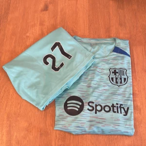 FC Barcelona bortatröja Lamine Yamal #27 - Snygg ljusblå FC Barcelona bortatröja med Lamine Yamal och nummer 27 på ryggen. Tröjan har klubbmärke, sponsorlogga (Spotify) och diskreta ränder i flera färger. Perfekt för dig som älskar Barca och vill sticka ut på läktaren.
