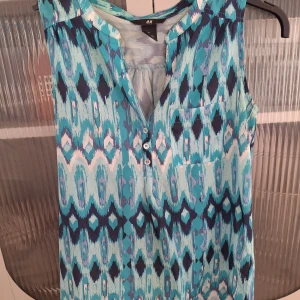 Mönstrad ärmlös blus från H&M - Säljer en ärmlös blus från H&M i storlek 34. Blusen har ett blått, turkost och svart mönster, knappar framtill och en liten bröstficka. Perfekt för varma dagar och enkel att matcha med jeans eller kjol.