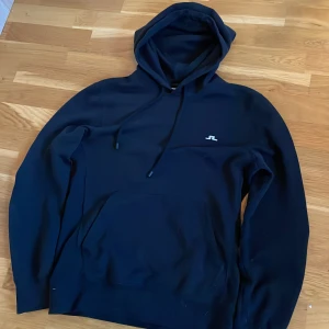 Svart hoodie från J.Lindeberg - Säljer en svart hoodie från J.Lindeberg med liten vit logga på bröstet. Tröjan har huva med dragsko och en stor magficka framtill. Perfekt för en avslappnad stil. Passar XS/S, pris kan diskuteras