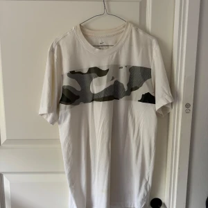 Vit t-shirt med camo-detalj från Nike - Säljer en vit t-shirt från Nike med en bred camouflagemönstrad panel över bröstet och stor Nike-logga. Klassisk passform och korta ärmar. Perfekt för en sportig och avslappnad stil.
