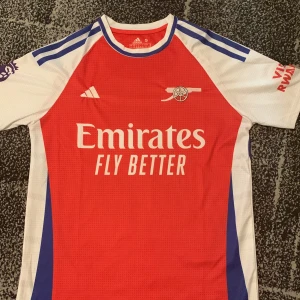 Arsenal fotbollströja  - Arsenal matchtröja från Adidas med Saka och nummer 7 på ryggen. Röd framsida med vita och blå detaljer, kortärmad modell. Emirates Fly Better-tryck på bröstet och klubbens emblem. Perfekt för fans av Arsenal och Saka.